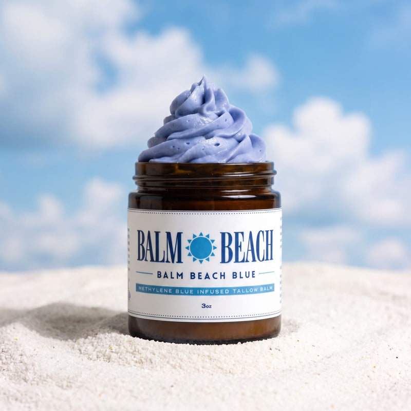 A jar of Balm Beach Blue moisturizing and infused body balm with a swirled dollop of the light blue balm on top, set on white sand with a blurred blue sky and clouds background.